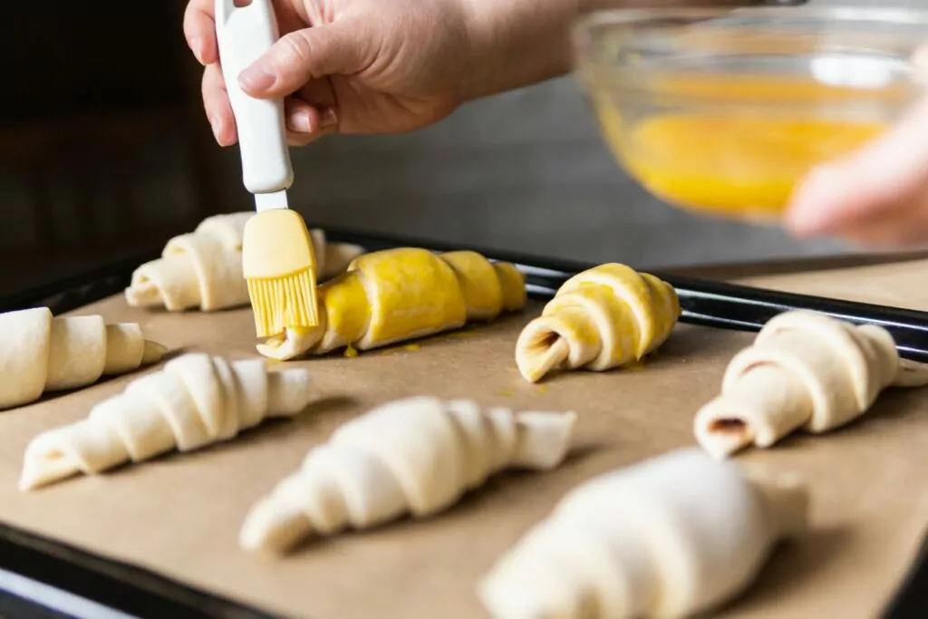 Baking croissants at home and covering the dough with egg yolk using pastry brush.