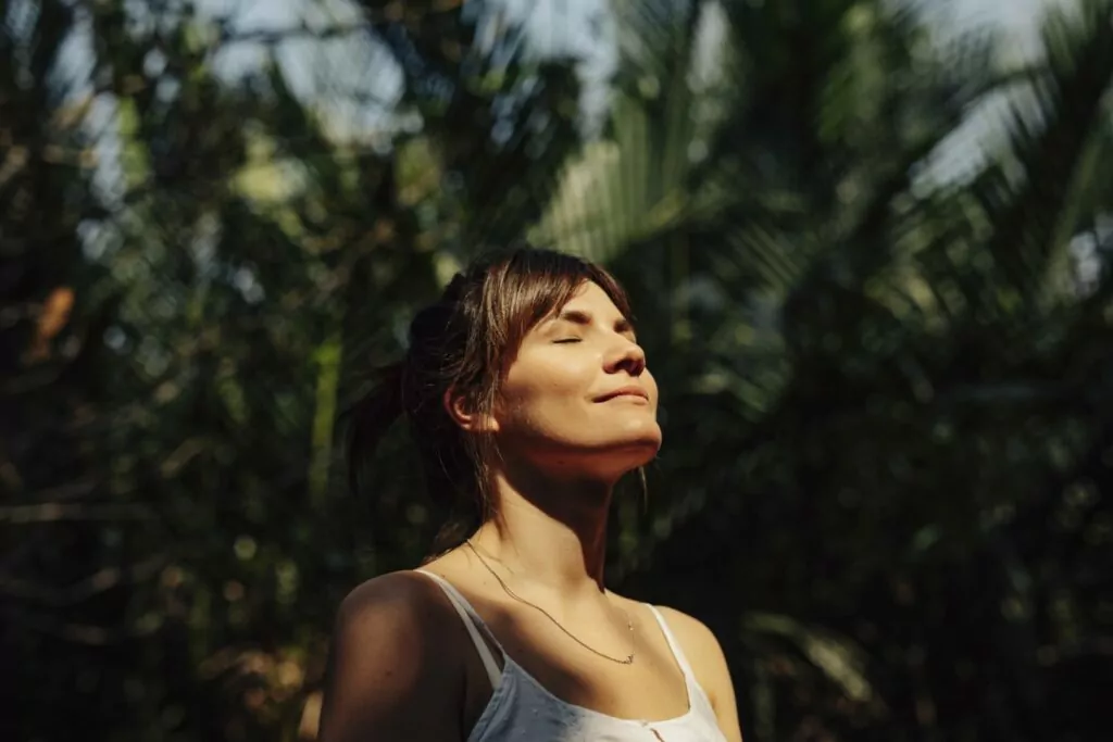 Close up upper body shot of a beautiful happy young Caucasian woman enjoying the warm sunlight and tropical atmosphere with her eyes closed surrounded by palm trees in a tropical public park.