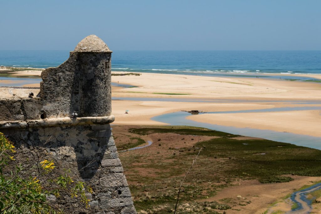 Tower of the old fortress made of stone. View on a sandy beach with puddles after a high tide. Horizon of the Atlantic ocean. Blue sky. Cacela Velha, Algarve, Portugal.