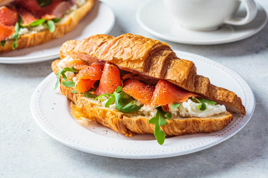 Croissant sandwich with cream cheese, salmon and arugula on a white plate, gray background, close-up. Healthy breakfast concept.