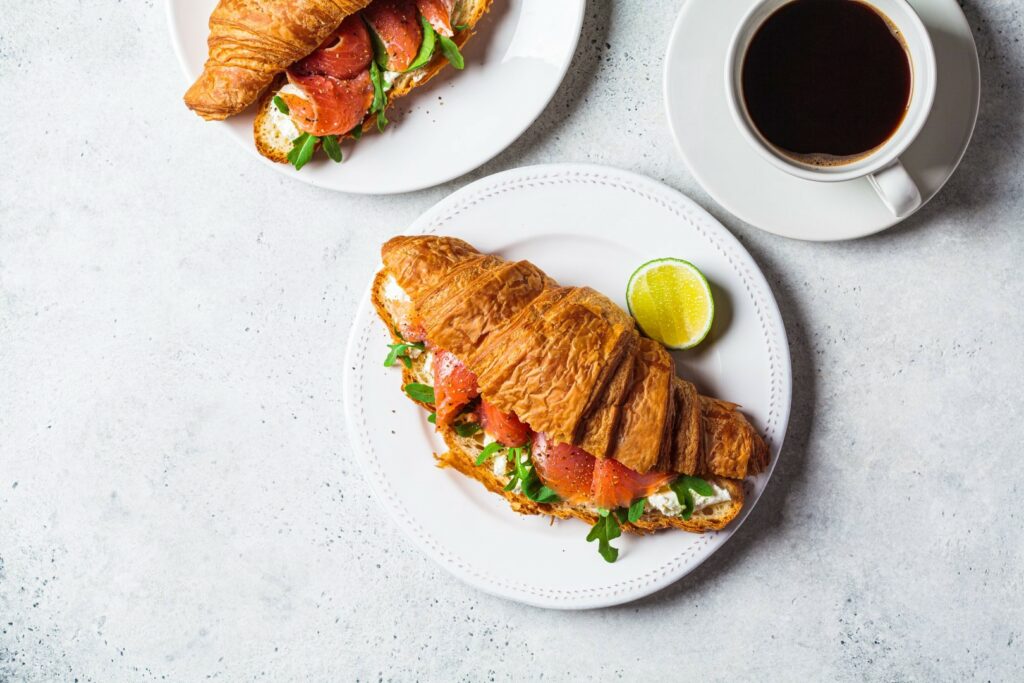 Croissant sandwich with cream cheese, salmon and arugula on a white plate, gray background, top view, copy space. Healthy breakfast concept.